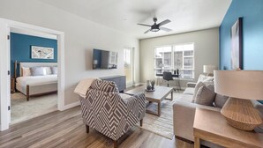Un salon moderne avec un canapé, un fauteuil et une table basse. On y trouve également un coin repas avec une table et des chaises, ainsi qu’une chambre avec un lit et un téléviseur.