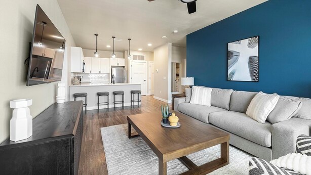 A modern living room with a grey sofa, a wooden coffee table, and a kitchen area with white cabinets and stainless steel appliances. - Landing | Amazing 2BD, Clubhouse (Goodyear)