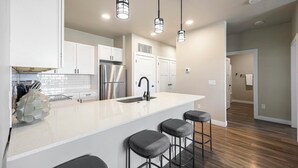 A modern kitchen with white cabinets, a stainless steel refrigerator, a sink, and bar stools. - Landing | Amazing 2BD, Clubhouse (Goodyear)