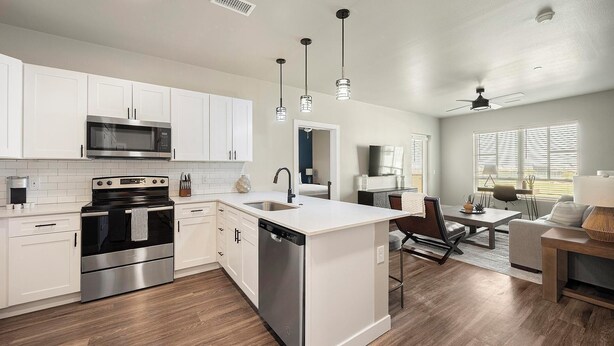 A modern kitchen with white cabinets, stainless steel appliances, and a central island. There is a dining area with a table and chairs, and a living room with a sofa and a TV. - Landing | Beautiful 2BD, Clubhouse (Goodyear)