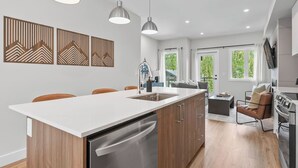 A modern kitchen with a central island, wooden cabinets, stainless steel appliances, pendant lighting, and a dining area with chairs. - Lumi | Slope-Side | Pvt Hot Tub | Resort Access (Panorama)