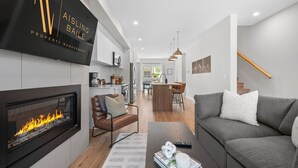 A modern living room with a fireplace, a sofa, a coffee table, and a kitchen in the background. - Lumi | Slope-Side | Pvt Hot Tub | Resort Access (Panorama)