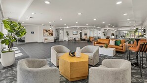 Een ruime lobby met grijs gestoffeerde stoelen, een houten salontafel en een receptiehoek met een bank en een televisie.
