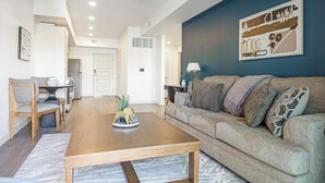A living room with a wooden coffee table, a grey sofa with patterned cushions, and a dining area with a white table and chairs. - Landing | Chic 1BD, Gym, Pool (Phoenix)