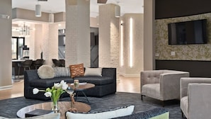 A modern living room with a dark grey sofa, a round coffee table, and a grey armchair. There is a large TV mounted on the wall and a staircase in the background.