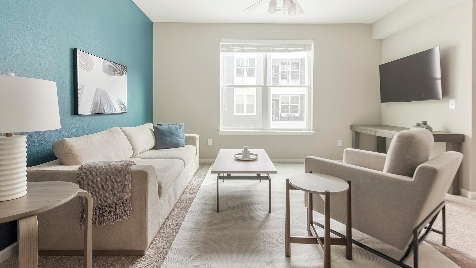 Una sala de estar con sofá, mesa de centro y sillón, un televisor montado en la pared, y una ventana con persianas.
