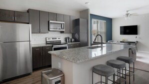 Une cuisine moderne avec des meubles de couleur sombre, des appareils en acier inoxydable, un îlot central doté de tabourets de bar et un téléviseur à écran plat fixé au mur.