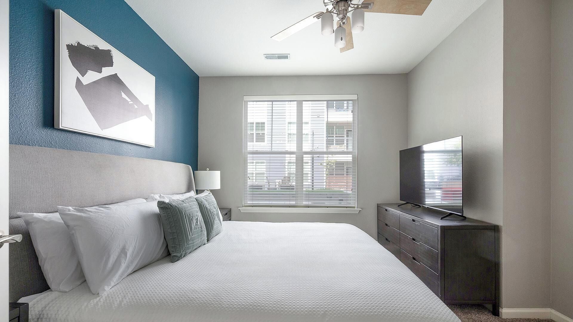 Une chambre à coucher avec un lit, un téléviseur, un ventilateur de plafond, une fenêtre et des œuvres d’art accrochées au mur.