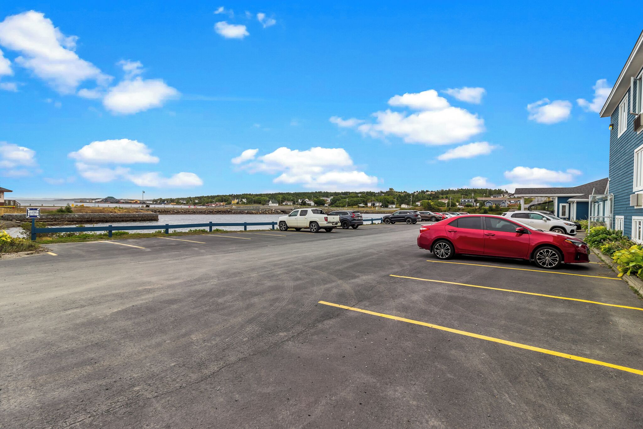 A parking lot with several cars, a blue building, and a clear blue sky with clouds.