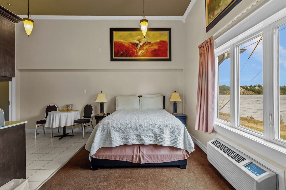 A bedroom with a bed, bedside lamps, a table with chairs, a painting, and a window with a view.