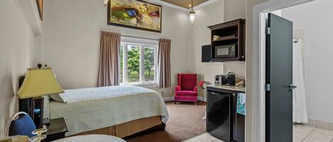 A hotel room with a bed, a desk with a microwave, a red chair, and a painting on the wall.