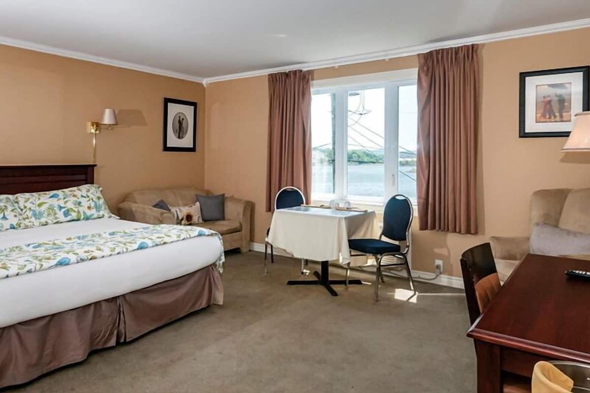 A hotel room with a bed, a sofa, a dining table with chairs, and a view of a body of water through large windows.
