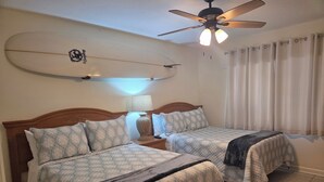 A hotel room with two beds, a ceiling fan, a nightstand with a lamp, and a surfboard on the wall. - Unit # 317 (Cocoa Beach)