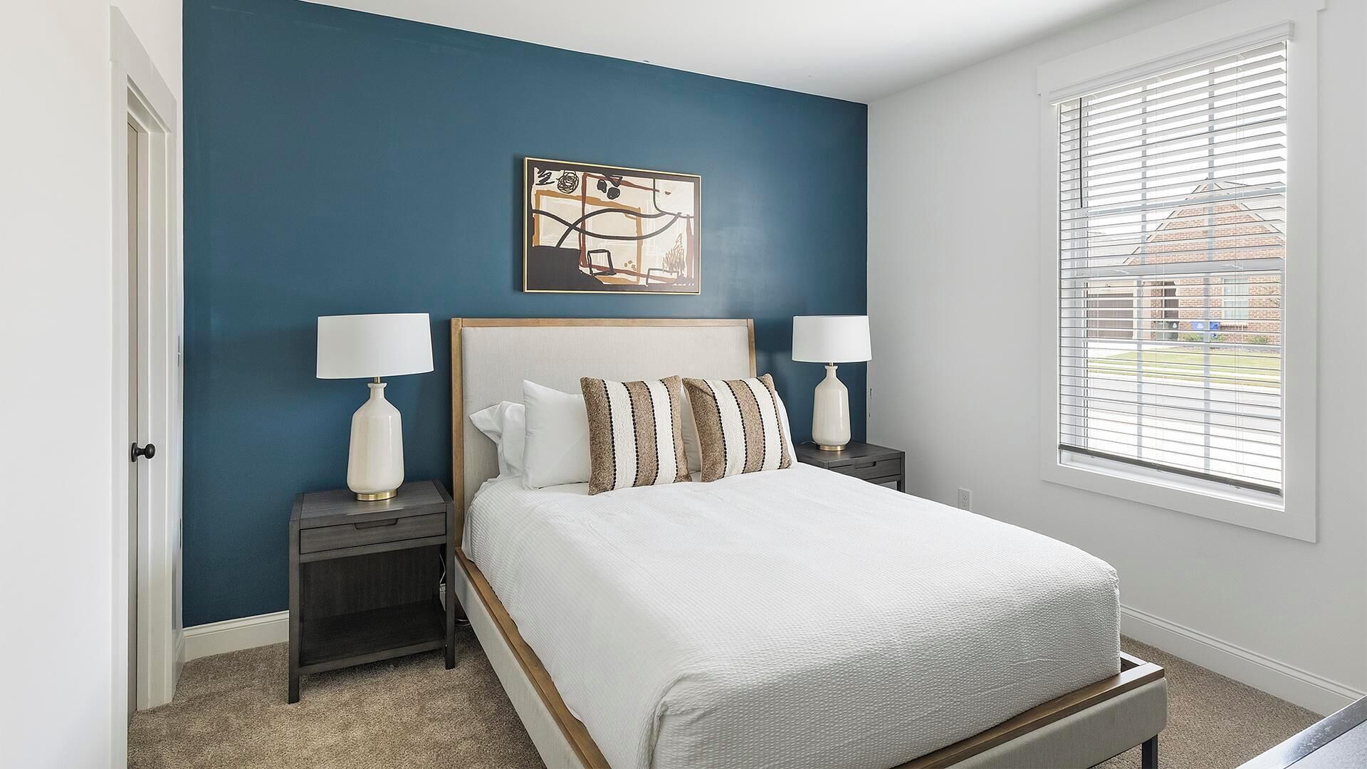 A bedroom with a bed, two bedside tables, a window with blinds, and a wall with a painting.