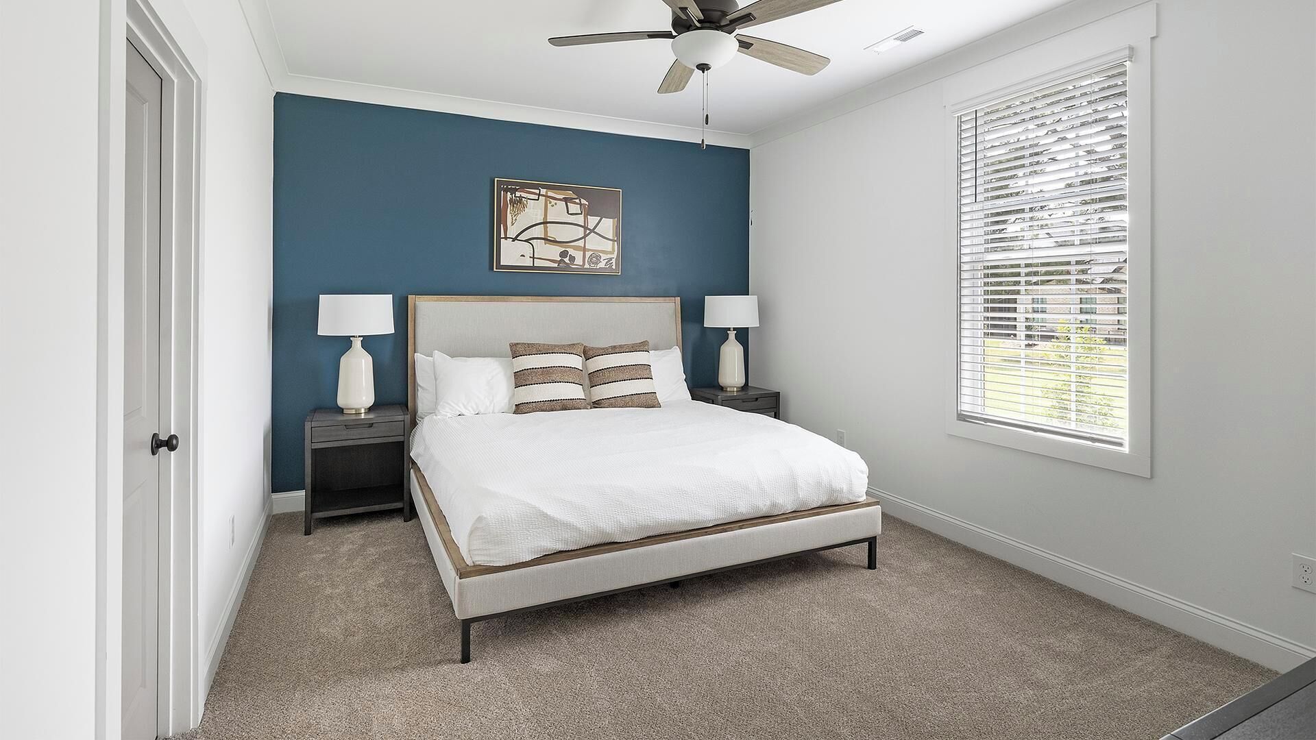 A bedroom with a bed, two bedside tables, a ceiling fan, a window with blinds, and a wall-mounted artwork.