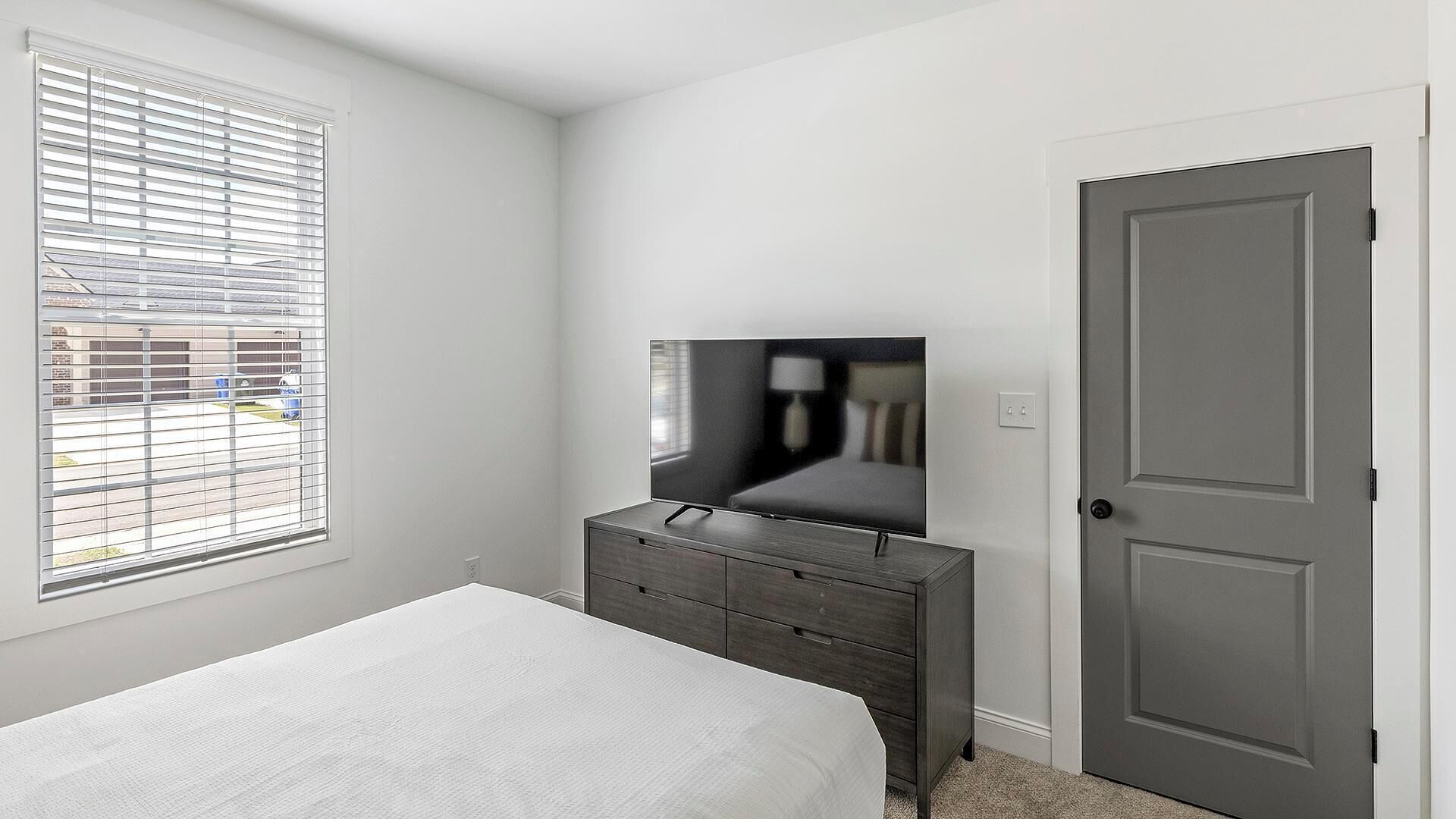 A bedroom with a bed, a dresser, a door, and a window with blinds.