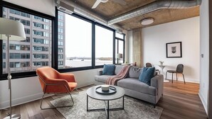 A modern living room with a large window, a grey sofa, an orange chair, a round coffee table, and a small side table.
