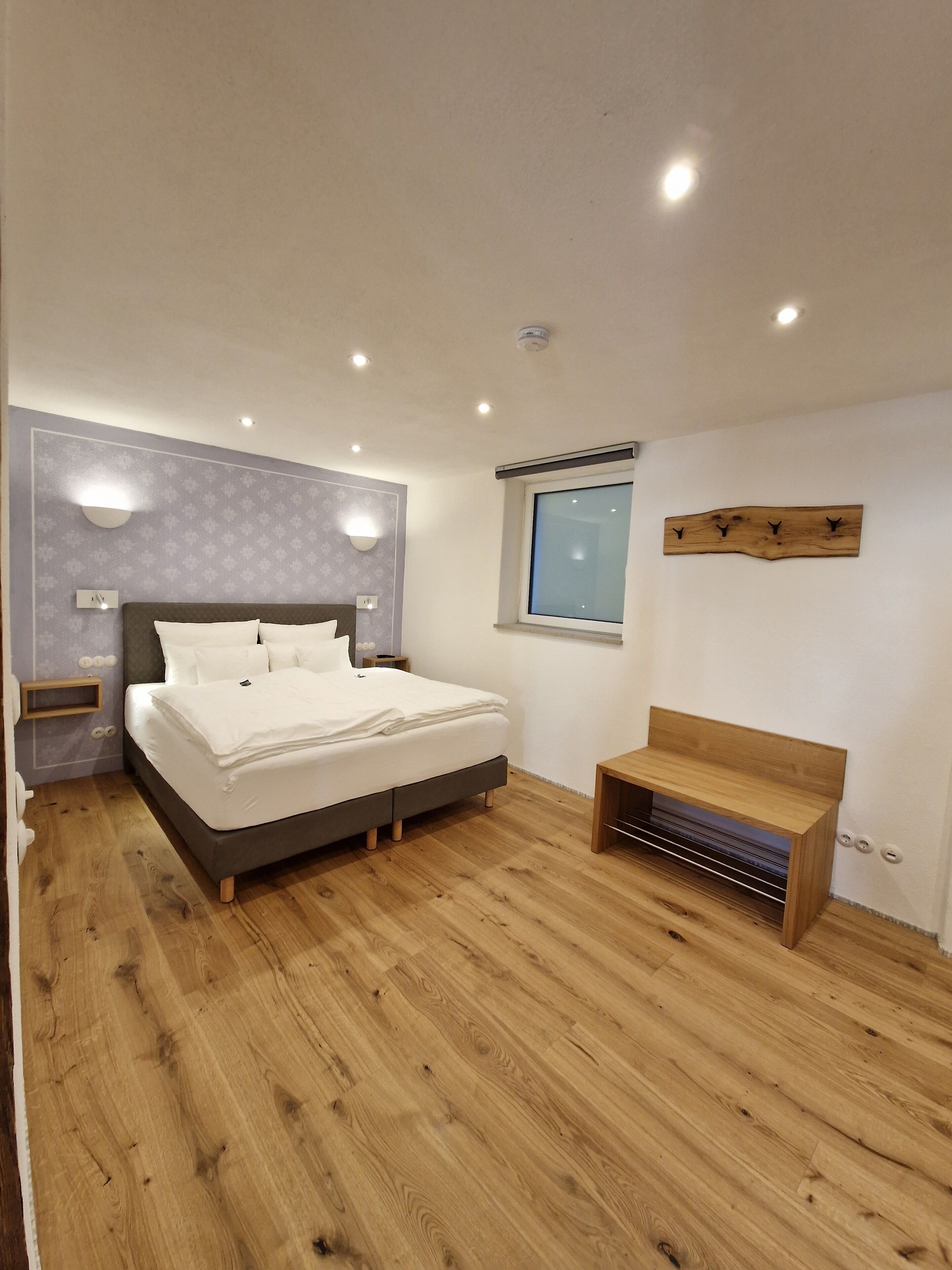 Une chambre à coucher comprenant un lit, des tables de chevet, un banc en bois, une fenêtre et une étagère fixée au mur.