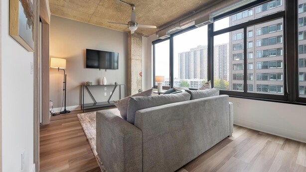 A modern living room with a grey sofa, a standing lamp, a flat-screen TV, and a large window offering a city view. - Landing | Spectacular 1BD, Gym, Game Room (Arlington)
