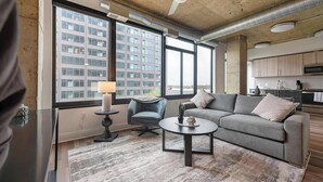 A modern living room with a grey sofa, a small round table, and a blue chair. There is a large window providing a view of a building outside. - Landing | Spectacular 1BD, Gym, Game Room (Arlington)