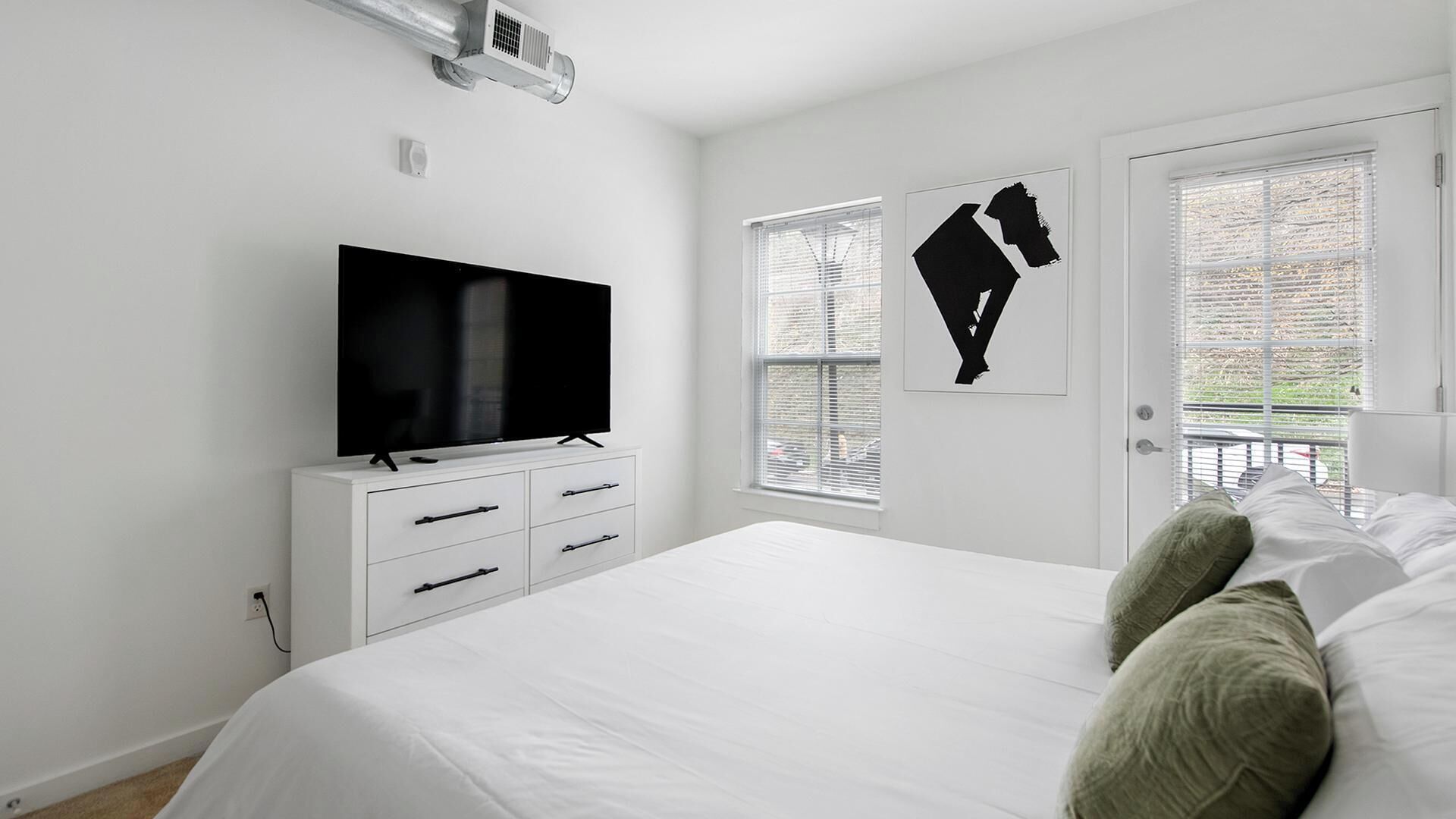 A bedroom with a large bed, a TV on a white dresser, a window with blinds, and a door with a black silhouette artwork.