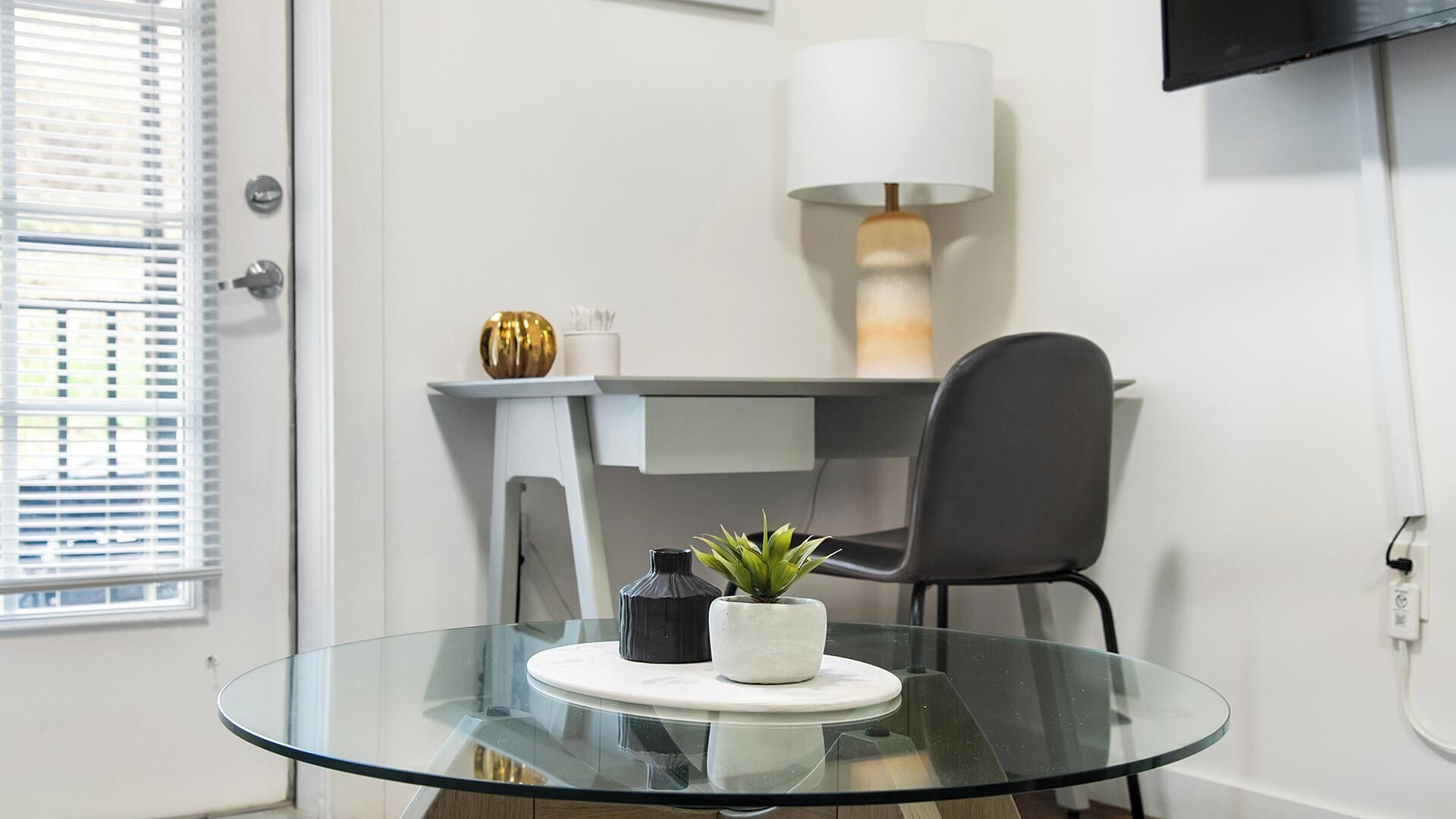 Een moderne kamer met een glazen tafel, een zwarte stoel, een witte lamp en een kleine plant op de tafel.