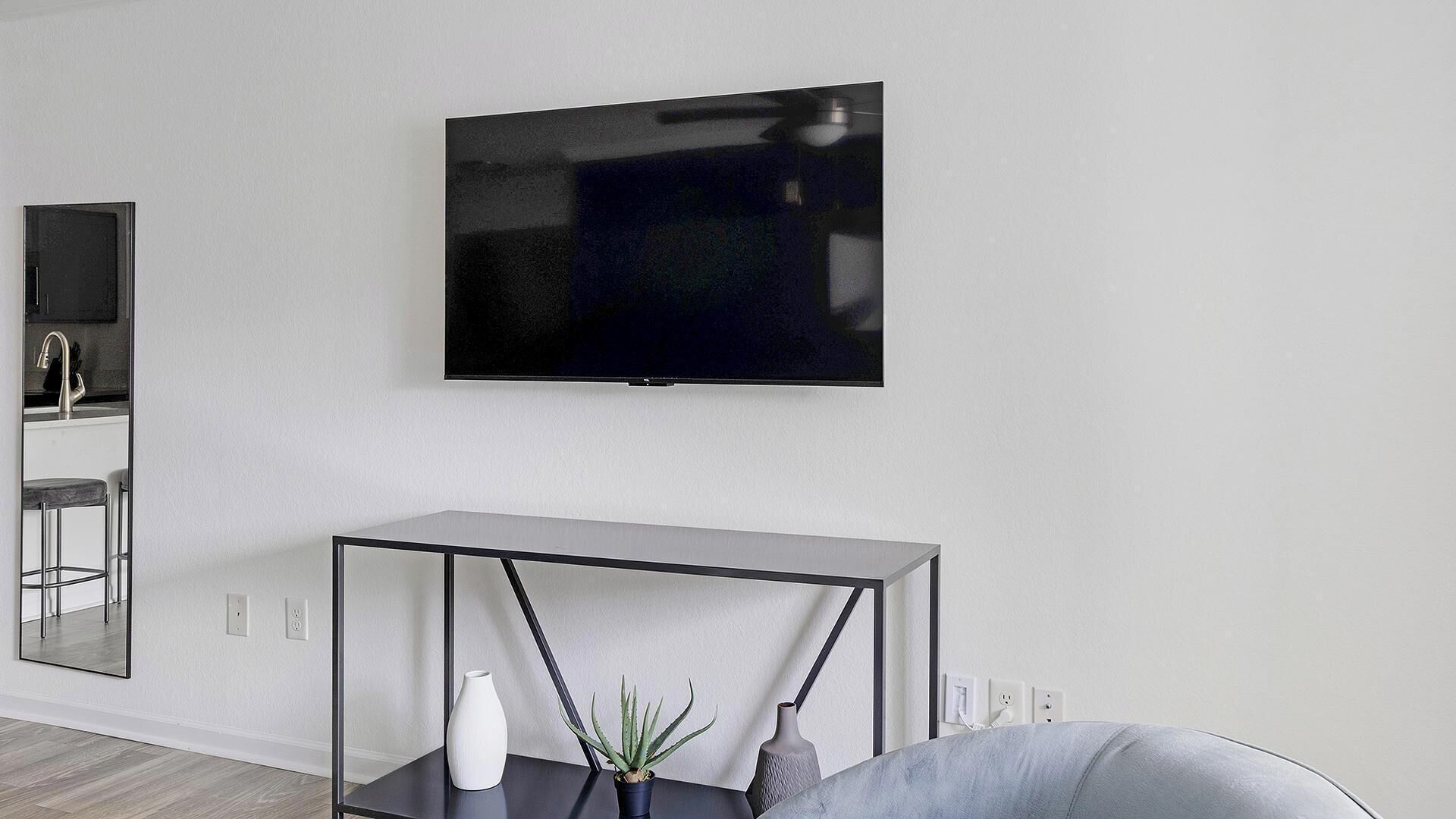 En moderne stue med et fladskærms-tv monteret på væggen, et sort konsolbord med en vase og plante, samt et stort spejl der reflekterer rummet.