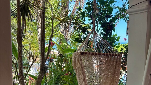 A hanging hammock in a lush, green outdoor area with a tiled floor and a white wall.
