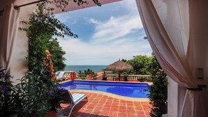 A pool area with a view of the ocean, surrounded by potted plants and a thatched umbrella.