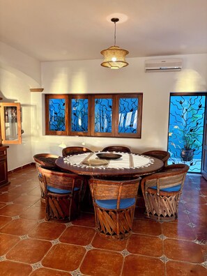 A dining area with a round table, wicker chairs, a woven ceiling light, and a view through a window with a blue wavy pattern.