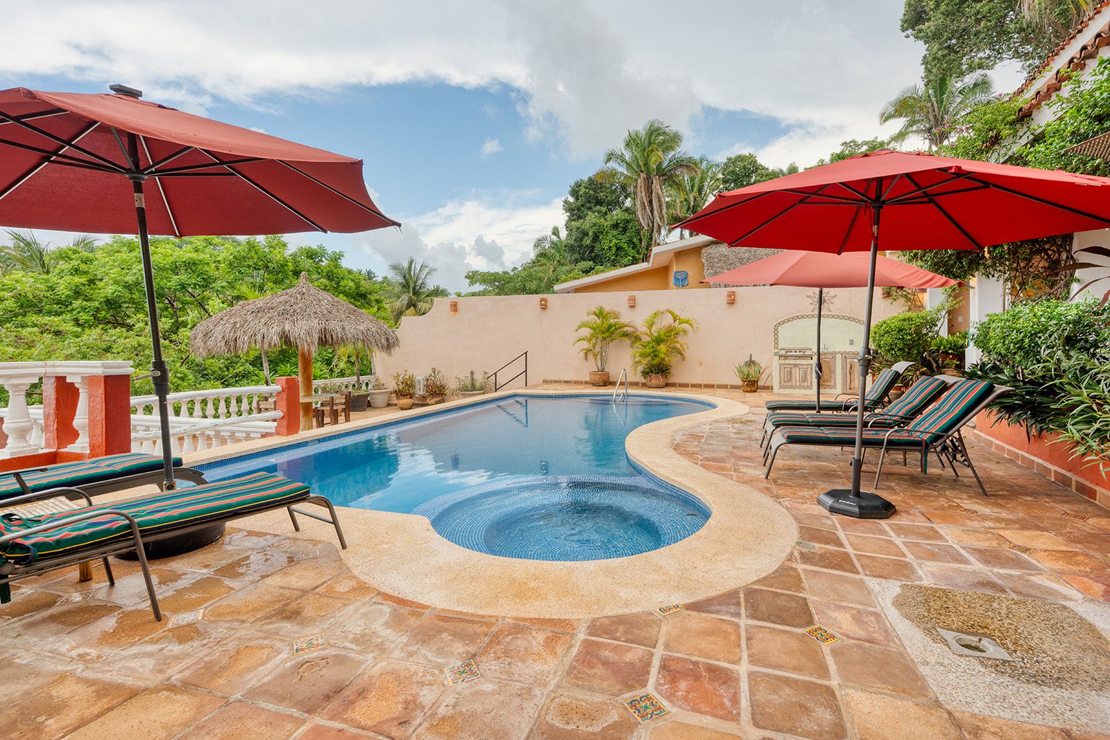 A swimming pool with a hot tub, surrounded by lounge chairs and umbrellas, set on a stone patio with a view of greenery and a building in the background.