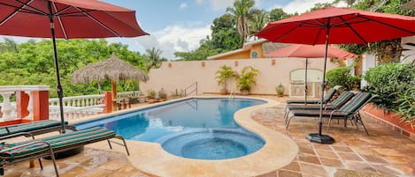 A swimming pool with a hot tub, surrounded by lounge chairs and umbrellas, set on a stone patio with a view of greenery and a building in the background.