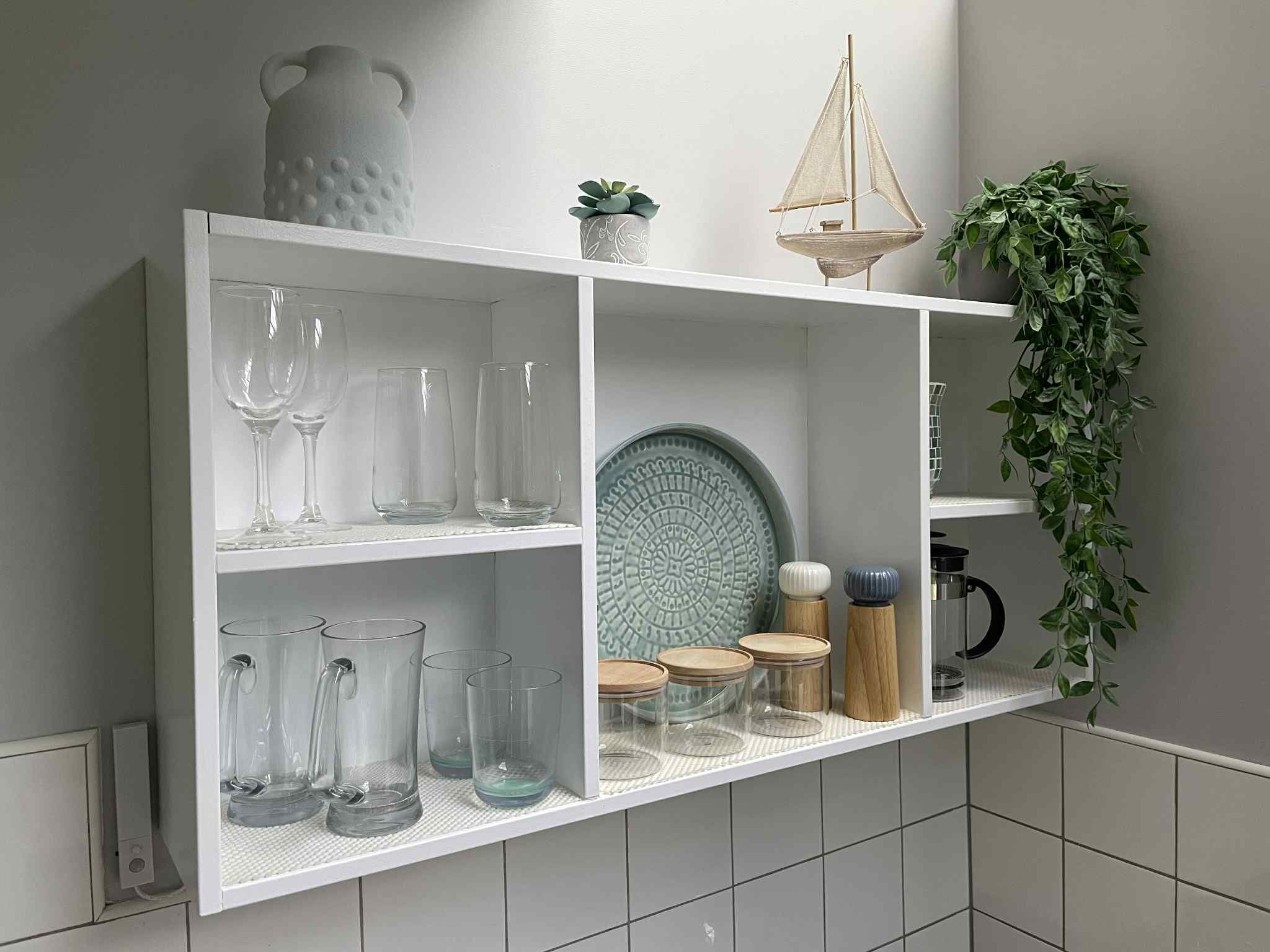 A white shelf unit with various glassware, a decorative plate, and a hanging plant.