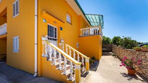 A yellow building with a staircase, a balcony, and a stone wall with potted plants.