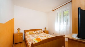 A bedroom with a wooden bed, two bedside tables with lamps, a wooden desk with a television, and a window with sheer curtains.