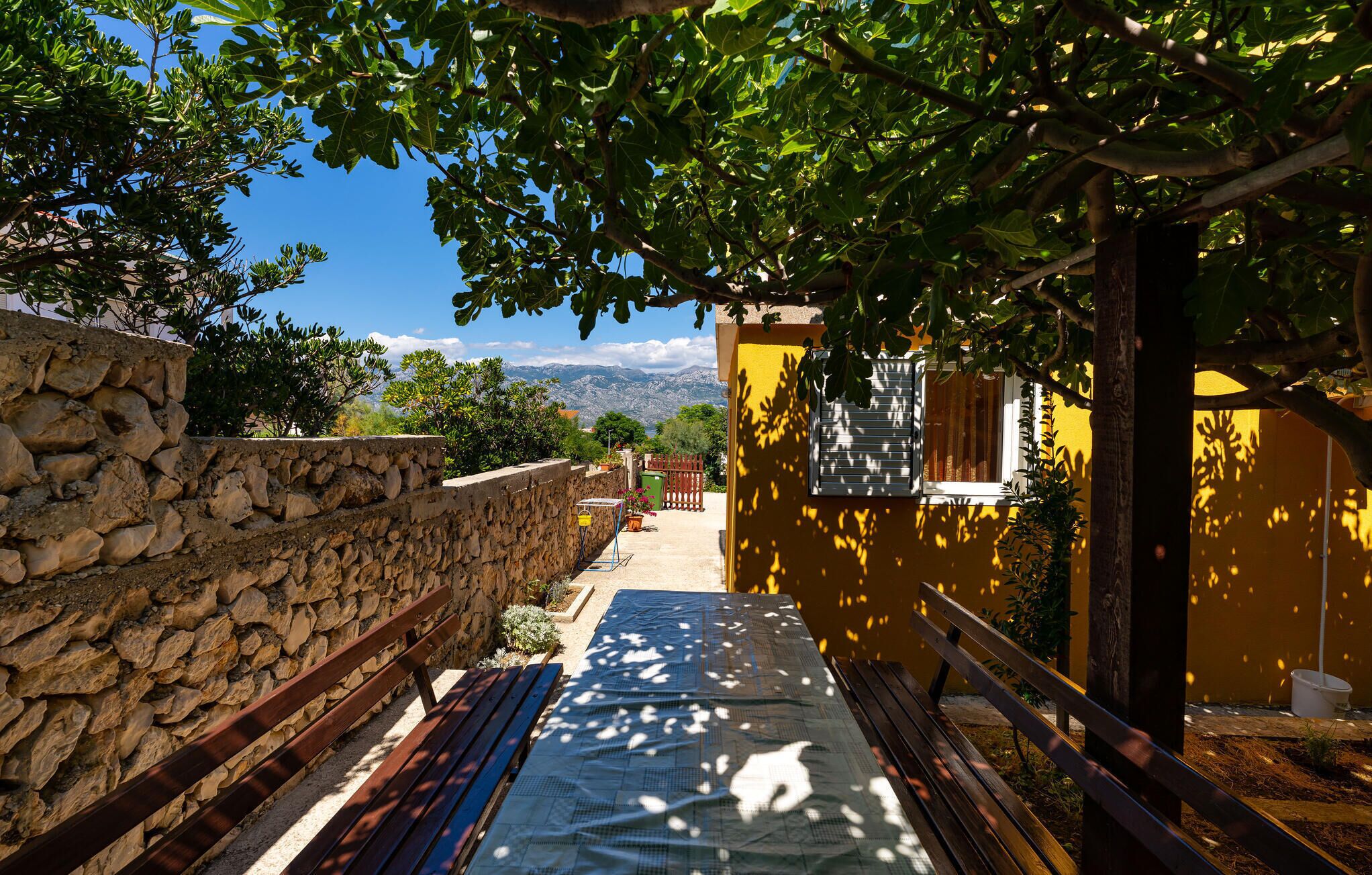 A stone wall, a wooden bench, a yellow building, and a view of mountains.