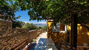A stone wall, a wooden bench, a yellow building, and a view of mountains.