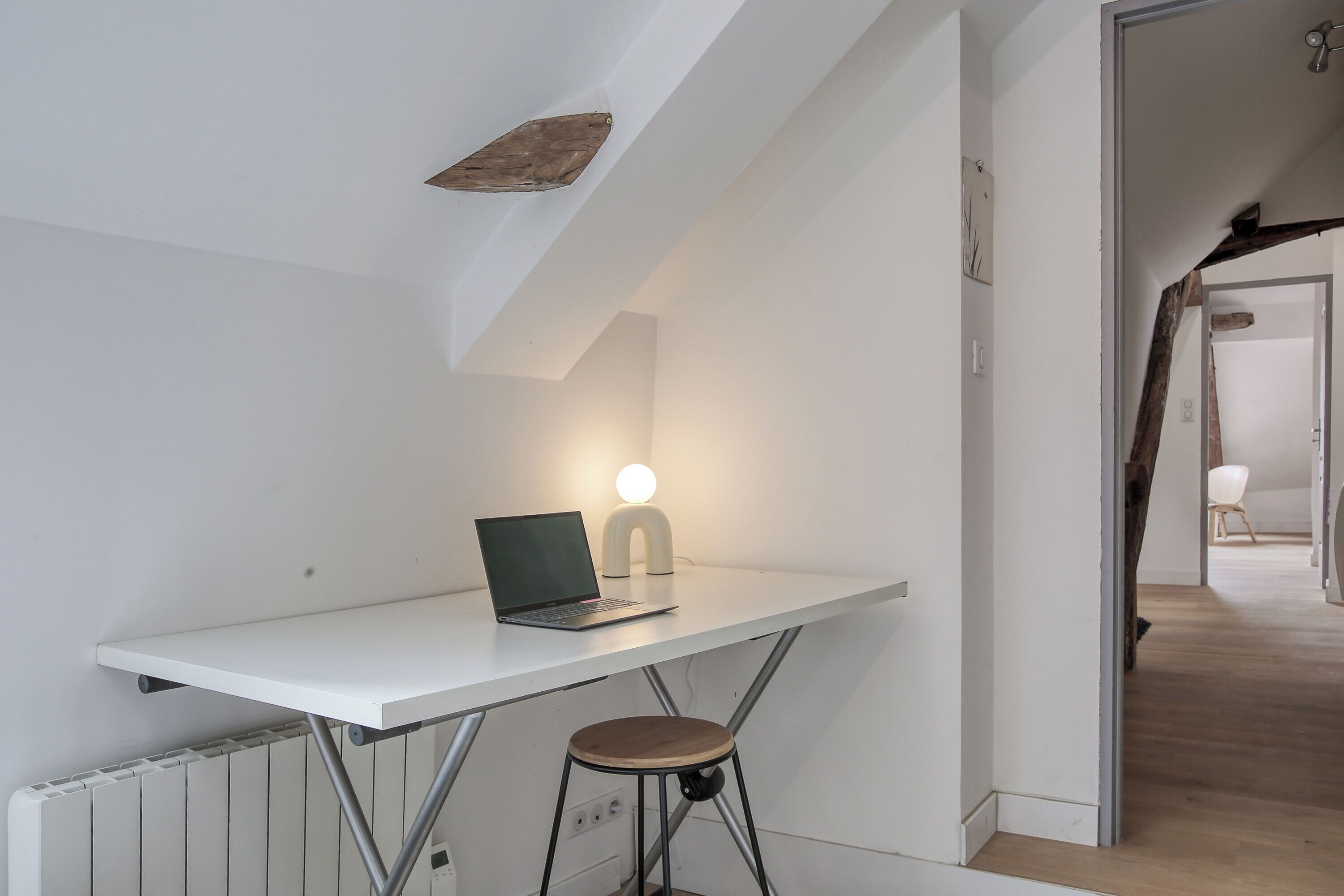 Een wit bureau met een laptop en een lamp in een kamer met witte muren en een schuin dak.