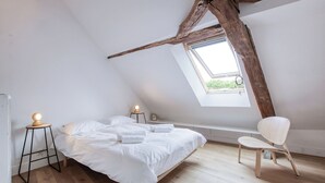 A bedroom with a sloped ceiling, a bed with white linens, a wooden chair, and a small table with a lamp.