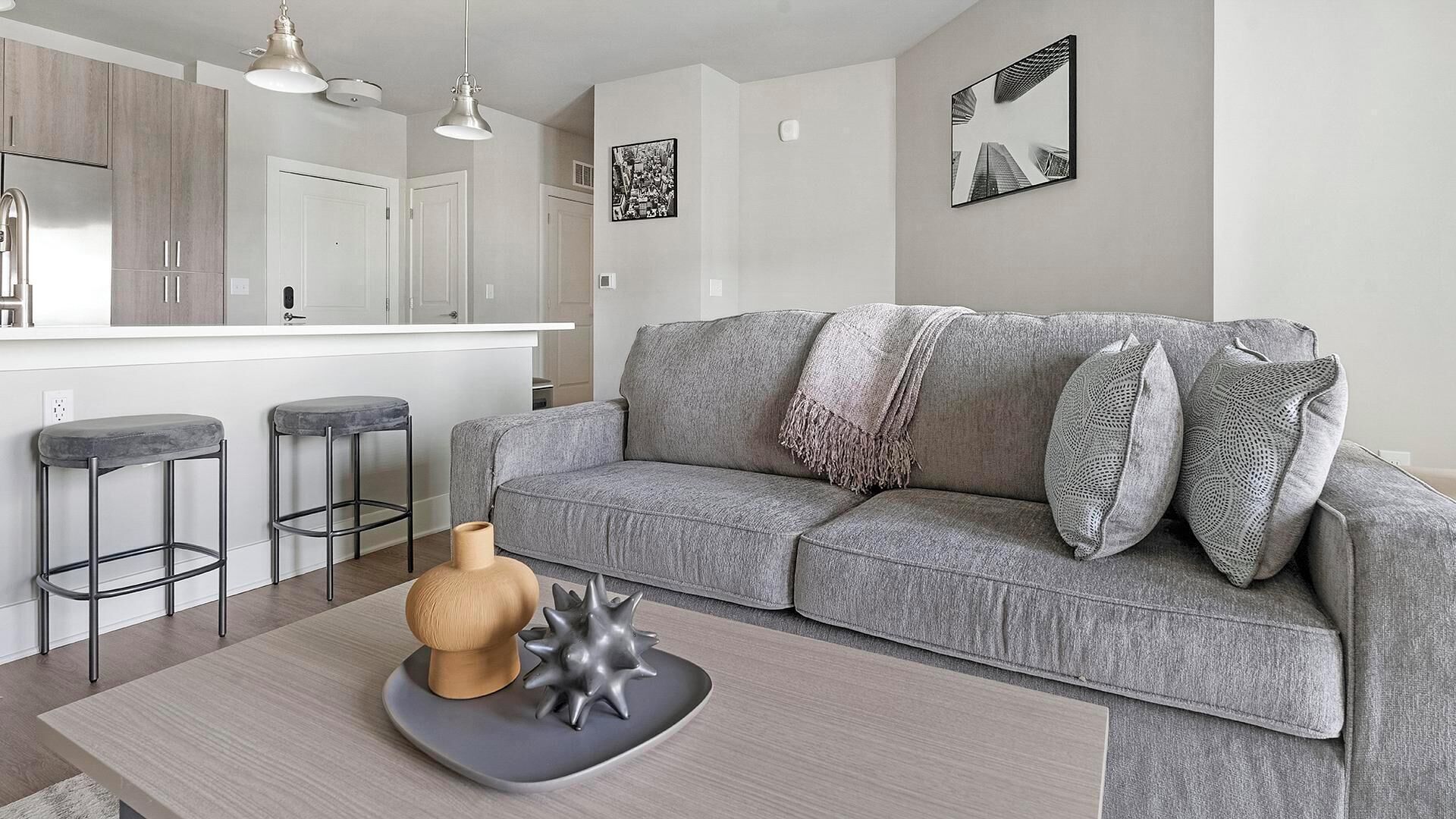 A modern living room with a grey sofa, two bar stools, a coffee table with decorative items, a kitchen area with white cabinets, and artwork on the walls.