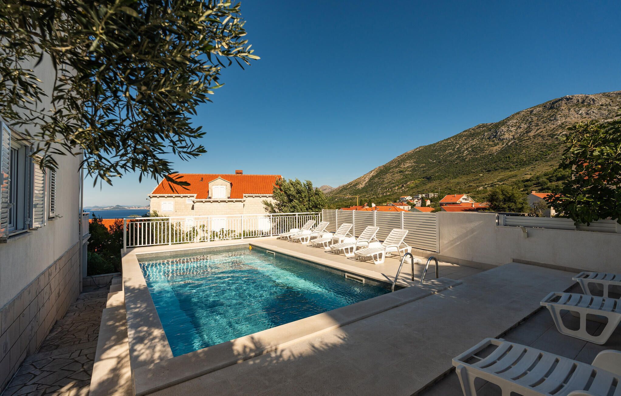 Ein Pool mit Liegestühlen, Blick auf ein Gebäude mit rotem Dach und eine gebirgige Landschaft im Hintergrund.