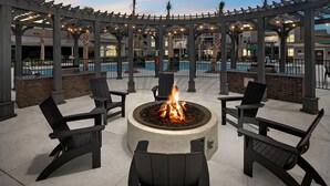 Ein Außenbereich mit einer runden Feuerstelle, umgeben von Sitzgelegenheiten, mit einem Pool im Hintergrund und einer überdachten Pergola.