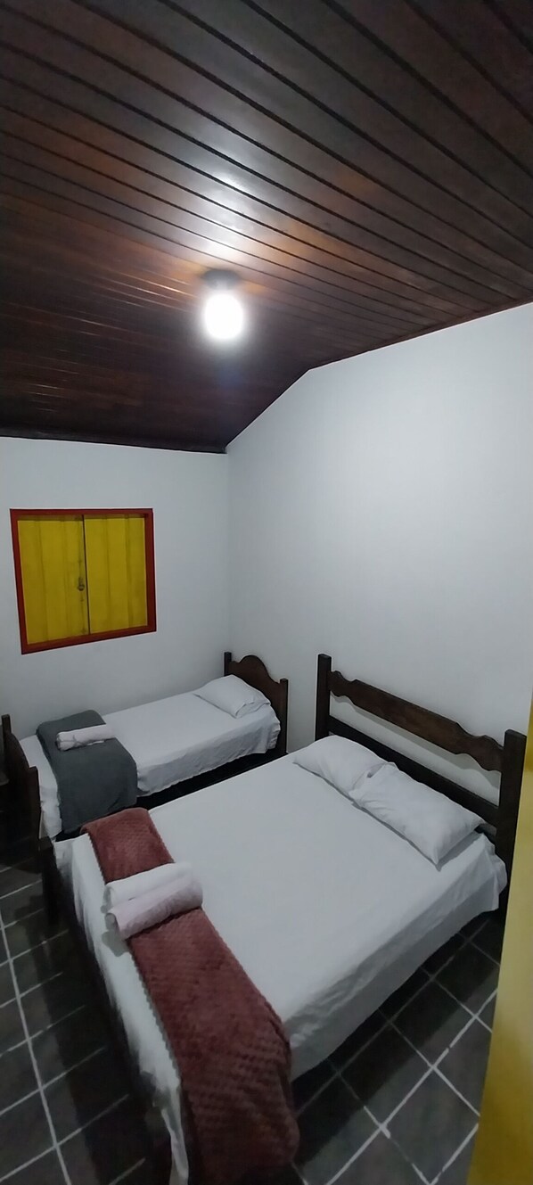 A room with two single beds, a wooden ceiling, a yellow-framed window, and a tiled floor.