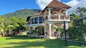 Une maison à deux étages dotée d’un balcon, entourée d’une pelouse bien entretenue et d’une végétation tropicale.