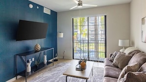 A modern living room with a blue accent wall, a flat-screen TV, a ceiling fan, a sofa, and a large window with a view of buildings and greenery.