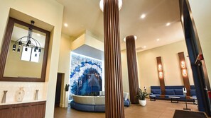 A hotel lobby with a reception desk, a large wall mural, a blue sofa, and tall wooden columns. - Landing | Spectacular 2BD, Pool, Gym (Fort Lauderdale)