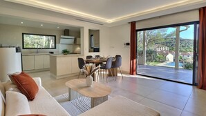 Sala moderna com sofá, mesa de centro e área de jantar. Uma cozinha é visível ao fundo, e uma grande porta de vidro deslizante dá acesso a uma varanda com vista para uma casa e árvores.