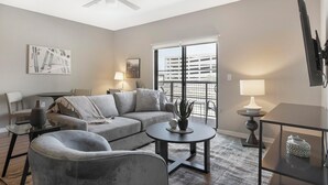 A modern living room with a grey sofa, two armchairs, a coffee table, and a dining table. There is a balcony with a view of a building. - Landing | Stylish 1BD, Gym, Game Room (Phoenix)
