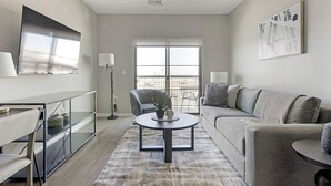 Ein modernes Wohnzimmer mit Sofa, Couchtisch, einem an der Wand montierten Fernseher und einem großen Fenster mit Blick ins Freie.