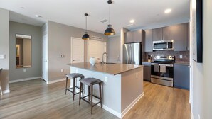 Une cuisine moderne dotée d’un îlot central, d’appareils électroménagers en acier inoxydable et de luminaires suspendus.